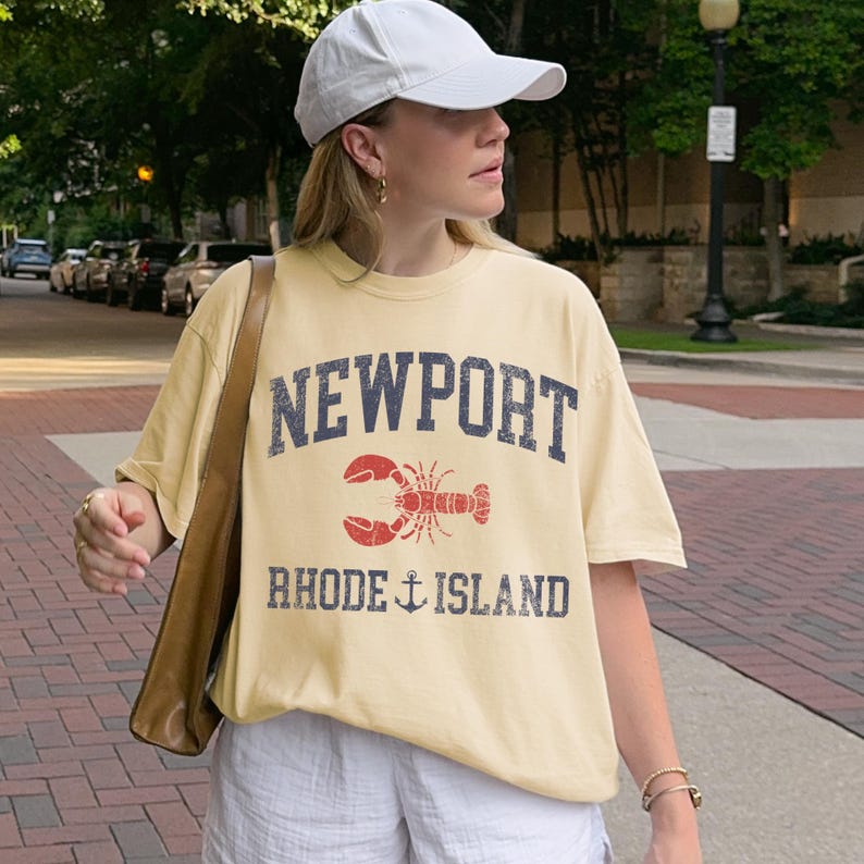 May include: A light yellow t-shirt with the word "NEWPORT" in navy blue block letters above a red lobster graphic. Below the lobster, the text "RHODE ISLAND" is printed in navy blue with an anchor symbol. The shirt is worn with a white baseball cap.