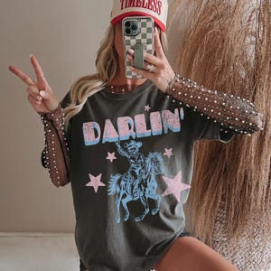 Puede incluir: Camiseta gris oscuro con la palabra "DARLIN'" en azul y rosa, con un vaquero montando a caballo. La imagen tambi&eacute;n muestra una gorra de b&eacute;isbol con la palabra "TIMELESS" y una funda de tel&eacute;fono.