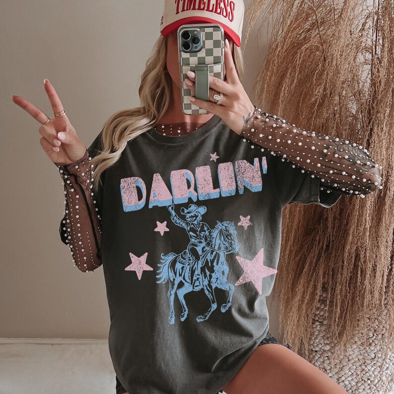 Puede incluir: Camiseta gris oscuro con la palabra "DARLIN'" en azul y rosa, con un vaquero montando a caballo. La imagen tambi&eacute;n muestra una gorra de b&eacute;isbol con la palabra "TIMELESS" y una funda de tel&eacute;fono.