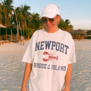 May include: White t-shirt with the word "NEWPORT" in blue, a red lobster graphic, and "RHODE ISLAND" with an anchor. The shirt is worn on a beach with palm trees in the background.