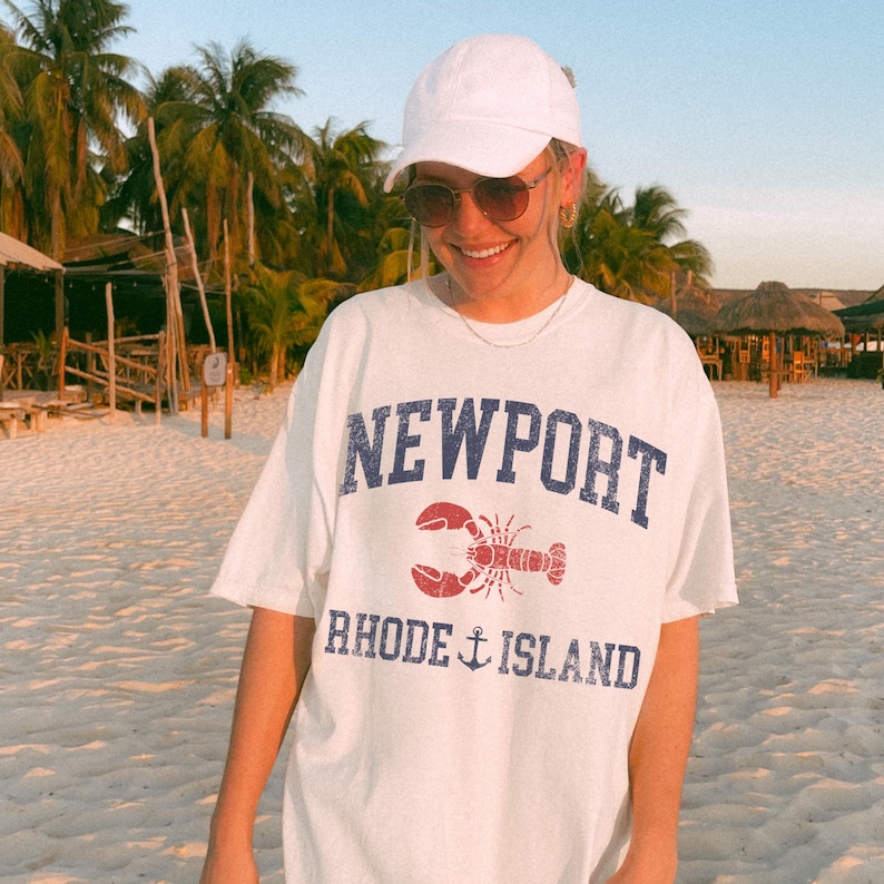 May include: White t-shirt with the word "NEWPORT" in blue, a red lobster graphic, and "RHODE ISLAND" with an anchor. The shirt is worn on a beach with palm trees in the background.