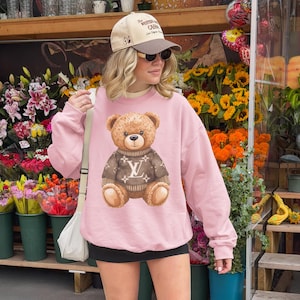 May include: A light pink sweatshirt featuring a brown teddy bear wearing a brown sweater with a white logo. The teddy bear has a friendly expression. The sweatshirt is a casual style, suitable for everyday wear.