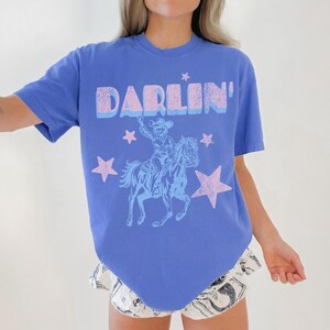 Puede incluir: Una camiseta azul b&iacute;garo con la palabra "DARLIN'" en una fuente en capas, azul claro y rosa. Debajo del texto hay una ilustraci&oacute;n azul claro de un vaquero montando a caballo, rodeado de estrellas rosas. La camiseta es de estilo casual y de manga corta.