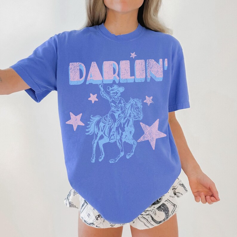 Puede incluir: Una camiseta azul b&iacute;garo con la palabra "DARLIN'" en una fuente en capas, azul claro y rosa. Debajo del texto hay una ilustraci&oacute;n azul claro de un vaquero montando a caballo, rodeado de estrellas rosas. La camiseta es de estilo casual y de manga corta.