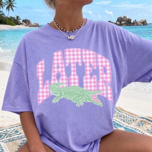 May include: A lilac-colored t-shirt featuring the word "LATER" in a pink and white gingham pattern. Below the text is a green alligator graphic with an open mouth. The shirt is a casual, oversized style.