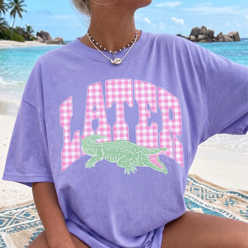 May include: A lilac-colored t-shirt featuring the word "LATER" in a pink and white gingham pattern. Below the text is a green alligator graphic with an open mouth. The shirt is a casual, oversized style.