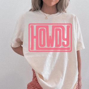 May include: A cream-coloured t-shirt featuring the word "HOWDY" in a pink rectangular design. The shirt has short sleeves and a relaxed fit. The model is wearing a gold necklace and a red and white checked skirt.
