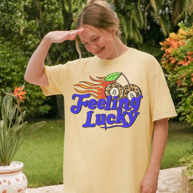 K&ouml;nnte beinhalten: Hellgelbes T-Shirt mit einem Aufdruck "Feeling Lucky". Das Design zeigt zwei Billardkugeln mit der Nummer 8, Flammen und ein gr&uuml;nes Blatt. Das T-Shirt ist ein l&auml;ssiger, bequemer Stil.