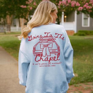 以下が含まれることがあります： 赤い車のグラフィックと「Going To The Chapel」と「& we're gonna get married!」の文字が入った、水色のスウェットシャツ。2025年も表示されています。スウェットシャツはカジュアルなスタイルです。