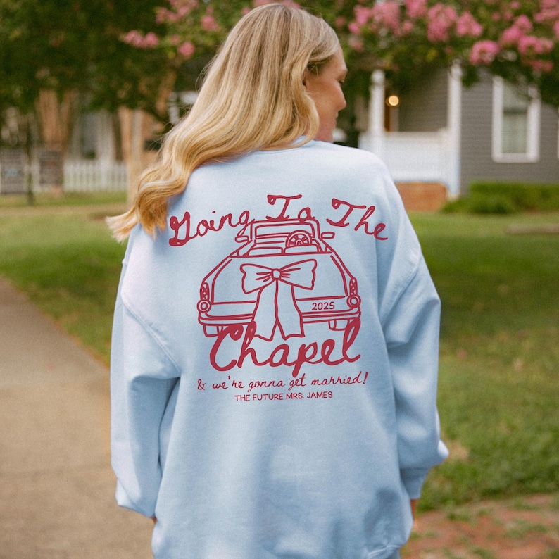 以下が含まれることがあります： 赤い車のグラフィックと「Going To The Chapel」と「& we're gonna get married!」の文字が入った、水色のスウェットシャツ。2025年も表示されています。スウェットシャツはカジュアルなスタイルです。