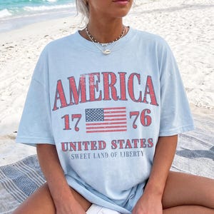 May include: Pale blue t-shirt featuring the word "AMERICA" in distressed red lettering. Below the word "AMERICA" is the number "1776" and a small American flag. The words "UNITED STATES" and "SWEET LAND OF LIBERTY" are printed below the flag.