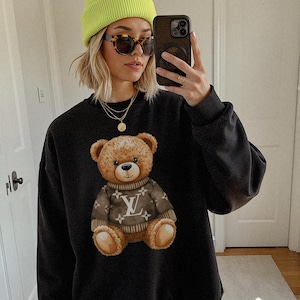 May include: Black sweatshirt featuring a brown teddy bear graphic wearing a brown sweater with a white logo. The sweatshirt has a crew neck and long sleeves. The person in the image is wearing a neon green beanie, sunglasses, and gold necklaces.