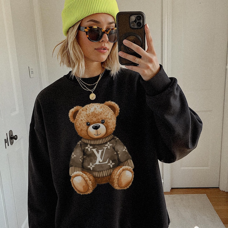 May include: Black sweatshirt featuring a brown teddy bear graphic wearing a brown sweater with a white logo. The sweatshirt has a crew neck and long sleeves. The person in the image is wearing a neon green beanie, sunglasses, and gold necklaces.