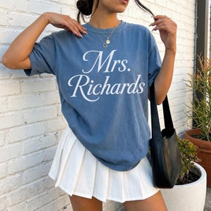 Może przedstawiać: Niebieski t-shirt z kr&oacute;tkim rękawem z białym napisem Mrs. Richards wydrukowanym kursywą z przodu. Koszulka jest zestawiona z białą plisowaną minisp&oacute;dniczką i czarną torbą na ramię, tworząc swobodny str&oacute;j uzupełniony delikatnym naszyjnikiem.