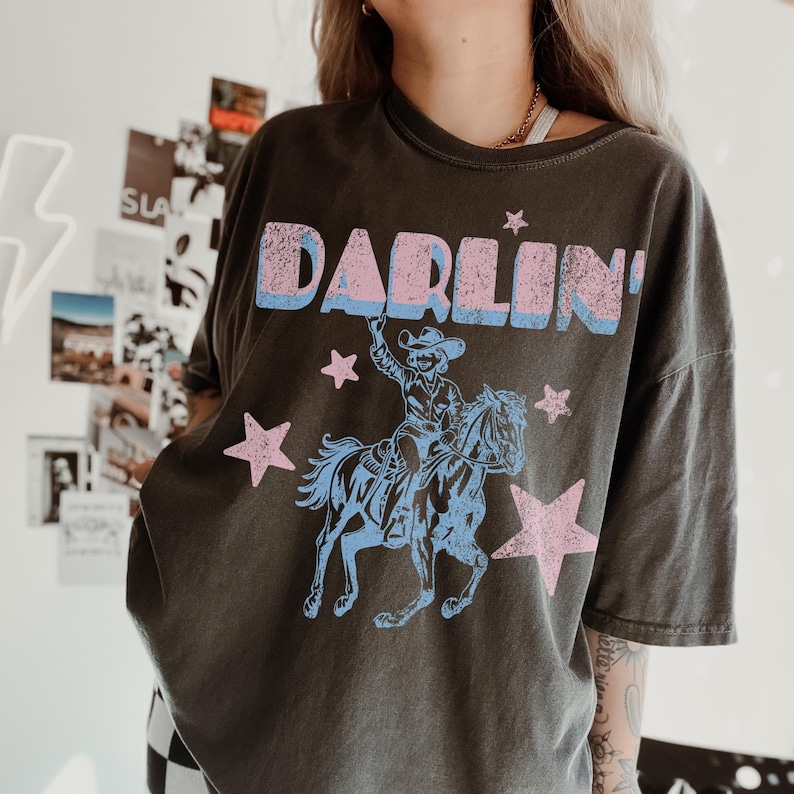 Puede incluir: Camiseta gris oscuro con la palabra "DARLIN'" en letras rosas y azules. Debajo, un gr&aacute;fico azul y rosa de un vaquero a caballo, con estrellas rosas. La camiseta tiene un aspecto vintage y desgastado, probablemente de algod&oacute;n.