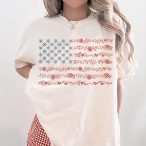 May include: Cream-colored t-shirt featuring a floral American flag design. The flag's stars are represented by blue floral patterns, and the stripes are composed of red floral vines. The shirt has short sleeves and a relaxed fit.