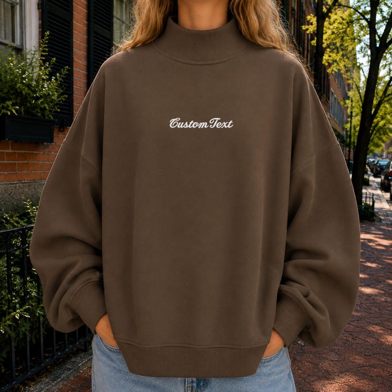 May include: A dark brown long-sleeve sweatshirt with a mock neck collar. The center of the chest features the text CustomText embroidered in a white cursive font. The garment has a relaxed fit with dropped shoulders and ribbed cuffs and hem.