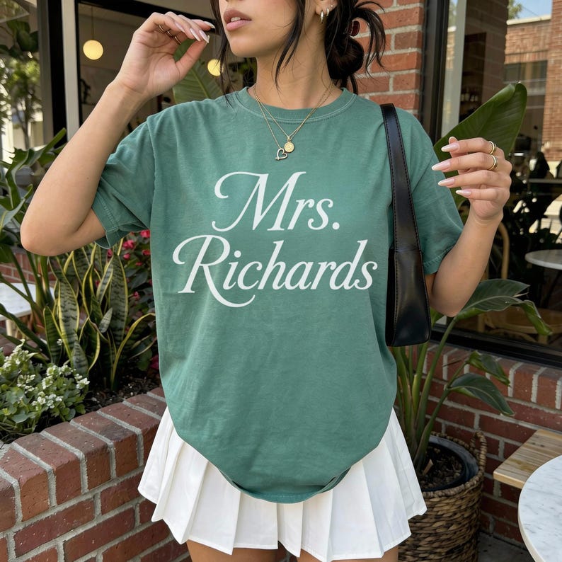 Może przedstawiać: Szałwiowo-zielony t-shirt z kr&oacute;tkim rękawem z białym napisem Mrs. Richards wydrukowanym kursywą na klatce piersiowej. Koszulka jest zestawiona z białą plisowaną sp&oacute;dnicą i czarną torbą na ramię, tworząc swobodny zestaw.
