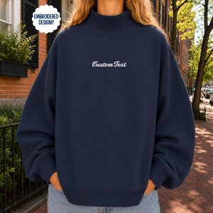 May include: A navy blue mock neck sweatshirt featuring long sleeves and a ribbed hem. The center of the chest displays the white embroidered text Custom Text. A circular white badge in the upper left corner contains the text EMBROIDERED DESIGN!!