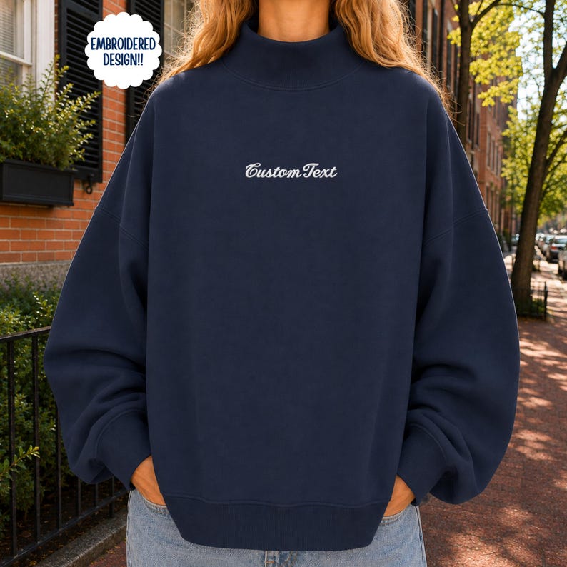 May include: A navy blue mock neck sweatshirt featuring long sleeves and a ribbed hem. The center of the chest displays the white embroidered text Custom Text. A circular white badge in the upper left corner contains the text EMBROIDERED DESIGN!!