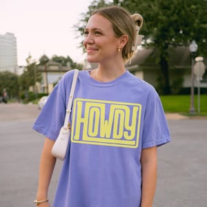 May include: A lilac t-shirt featuring the word "HOWDY" in a yellow, outlined font. The shirt has a loose fit and is being worn by a person. A white handbag with a gold clasp hangs from the person's shoulder.