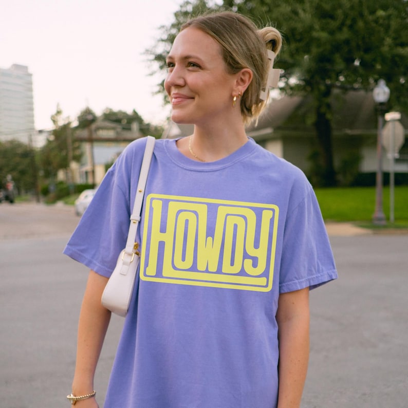 May include: A lilac t-shirt featuring the word "HOWDY" in a yellow, outlined font. The shirt has a loose fit and is being worn by a person. A white handbag with a gold clasp hangs from the person's shoulder.