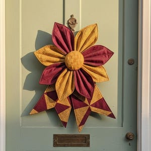 Pode incluir: Uma grande grinalda em forma de flor, feita à mão, em tons de bordô e dourado. As pétalas são em camadas, com um centro tecido. A grinalda está presa a uma porta verde clara com uma aldrava e uma ranhura para cartas de latão.