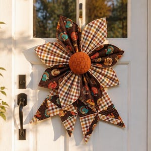 Peut inclure: Une décoration de porte en tissu en forme de grande fleur aux pétales alternés. Certains pétales présentent un fond marron avec un imprimé de dindes colorées, tandis que d'autres ont un motif à carreaux marron, blanc et jaune. Un centre orange texturé complète l'ensemble.