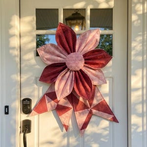 May include: A large, handmade fabric flower door decoration. The flower has alternating petals of solid and polka-dotted burgundy and light pink fabric. The center is a woven circle of pink fabric, and the bow is made of geometric shapes in matching fabrics.