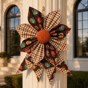 May include: A fabric wall hanging shaped like a flower with alternating petals featuring a brown background with colorful turkey prints and a brown, white, and orange plaid pattern. The center is a round, woven orange fabric button.