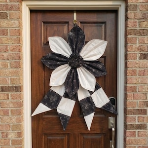 May include: A large, decorative fabric flower wreath in black and white hangs on a dark brown door. The flower has alternating white and black patterned petals, with a checkered pattern on the lower portion.