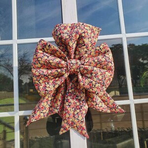 Może przedstawiać: Duża, dekoracyjna kokarda wykonana z kwiecistej tkaniny, z wzorem małych czerwonych, żółtych i białych kwiatów na ciemnym tle. Kokarda jest przymocowana do białej ramy okiennej, tworząc uroczy, vintage wygląd.