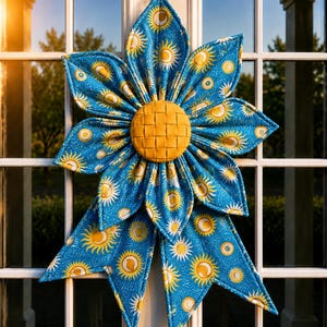 May include: A fabric wall hanging shaped like a large flower with blue petals featuring a yellow sun and moon pattern. The center of the flower is a round, woven yellow fabric piece. The petals are quilted and pointed, creating a star-like shape.