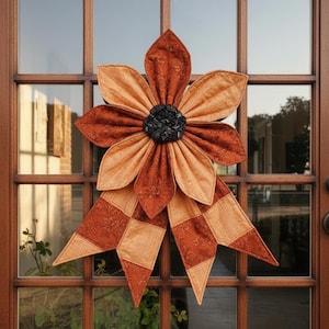 May include: A large, decorative flower made of fabric, featuring alternating shades of orange and tan. The center of the flower is a dark, textured material. The flower is attached to a door, creating a welcoming autumn-themed decoration.