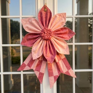 May include: A large, handmade fabric flower decoration in shades of pink and rose. The flower has layered petals and a textured center, with a checkered bow at the bottom. A small gold sun-shaped charm hangs from the top.