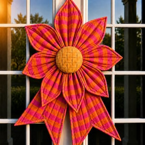 Op de afbeelding: Een stoffen wandhanger in de vorm van een bloem met doorgestikte bloemblaadjes in een felroze en oranje gestreept patroon. Het midden van de bloem is een ronde, gevlochten goudgele schijf. Het ontwerp bevat twee lange, puntige stoffen uiteinden.