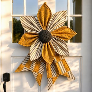 Pode incluir: Uma decoração de parede em forma de flor feita de tecido com pétalas alternadas em amarelo mostarda com um pequeno padrão circular preto e tecido branco com finas riscas verticais pretas. O centro é uma peça circular cinzento-escura.