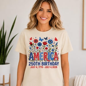 May include: A cream-coloured short-sleeve t-shirt featuring a graphic of red and blue flowers and butterflies. The text reads AMERICA 250TH BIRTHDAY JULY 4, 1776 - JULY 4, 2026. The word AMERICA is in a patriotic red, white, and blue polka dot pattern.