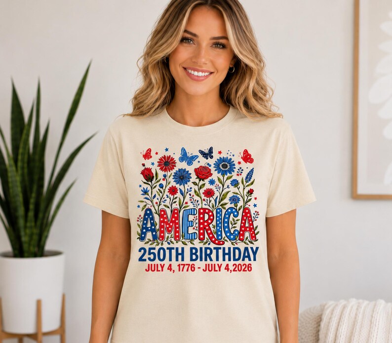 May include: A cream-coloured short-sleeve t-shirt featuring a graphic of red and blue flowers and butterflies. The text reads AMERICA 250TH BIRTHDAY JULY 4, 1776 - JULY 4, 2026. The word AMERICA is in a patriotic red, white, and blue polka dot pattern.