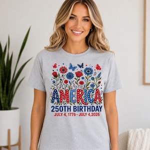 May include: A light grey short-sleeve t-shirt featuring a graphic of red and blue flowers, butterflies, and the text AMERICA 250TH BIRTHDAY JULY 4, 1776 - JULY 4, 2026. The word AMERICA is styled in red and blue with white star patterns.