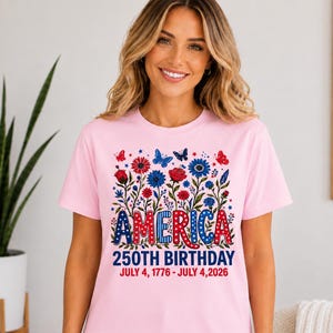 May include: A light pink short-sleeve t-shirt featuring a graphic of red and blue flowers, butterflies, and the text AMERICA 250TH BIRTHDAY JULY 4, 1776 - JULY 4, 2026. The design incorporates stars and polka dot patterns in red, white, and blue.