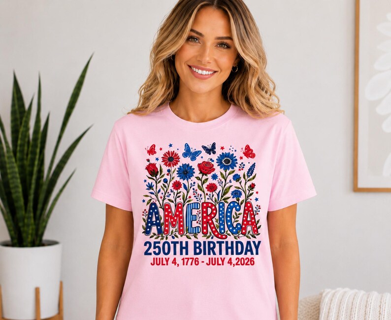 May include: A light pink short-sleeve t-shirt featuring a graphic of red and blue flowers, butterflies, and the text AMERICA 250TH BIRTHDAY JULY 4, 1776 - JULY 4, 2026. The design incorporates stars and polka dot patterns in red, white, and blue.
