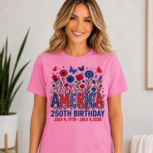 May include: A pink short-sleeved t-shirt featuring a graphic of red, white, and blue flowers and butterflies. The text reads AMERICA 250TH BIRTHDAY JULY 4, 1776 - JULY 4, 2026. The design uses a patriotic colour palette with stars and polka dot patterns.