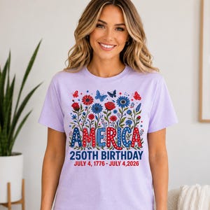 May include: A light purple t-shirt featuring a graphic of red and blue flowers and butterflies. The text reads AMERICA 250TH BIRTHDAY JULY 4, 1776 - JULY 4, 2026. The word AMERICA is styled in red, white, and blue patterns with stars and dots.