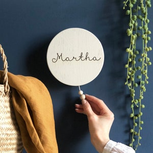 Peut inclure: Une pancarte ronde en bois blanc avec le nom Martha écrit en cursive noire. La pancarte est suspendue à une ficelle blanche avec deux perles en bois.