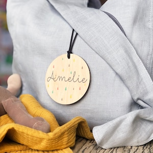 May include: A round wooden tag with the name "Amelie" written in cursive. The tag is decorated with colorful raindrops and has a black cord attached.