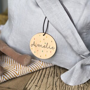 May include: A round wooden tag with the name "Amelie" written in cursive. The tag is decorated with colorful raindrops and is attached to a gray fabric bag.