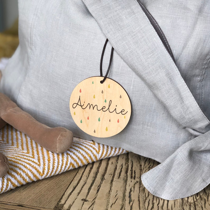 May include: A round wooden tag with the name "Amelie" written in cursive. The tag is decorated with colorful raindrops and is attached to a gray fabric bag.