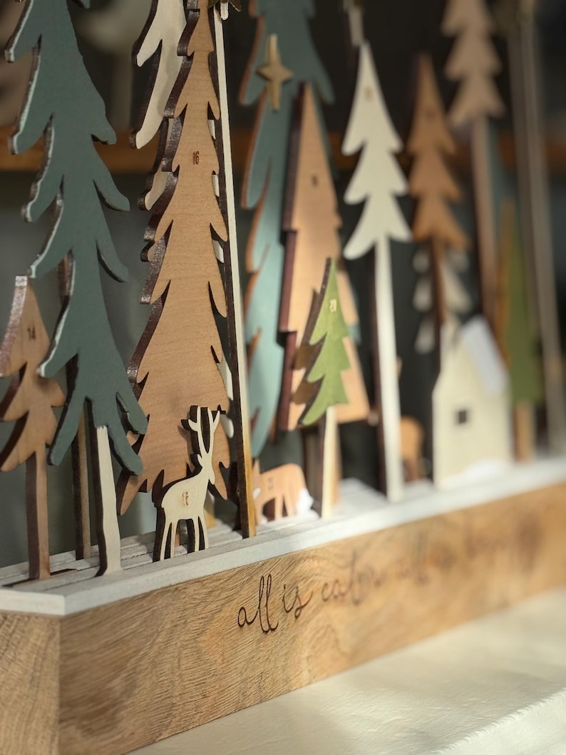 Pode incluir: Calend&aacute;rio do Advento em madeira com uma cena de floresta, incluindo &aacute;rvores, veados e uma casa. A base &eacute; gravada com "all is calm". As &aacute;rvores s&atilde;o verdes, castanhas e brancas. Decora&ccedil;&atilde;o de Natal.