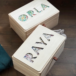 K&ouml;nnte beinhalten: Zwei Holzkisten mit ausgeschnittenen Buchstaben, die "ORLA" und "RAYA" auf den Deckeln bilden. Die ausgeschnittenen Buchstaben zeigen einen floralen Stoff. Jede Box hat eine kleine Lederlasche an der Seite.